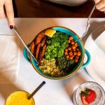 A vibrant vegan meal with fresh ingredients like broccoli, chickpeas, quinoa, served alongside a burrito and refreshing drinks.